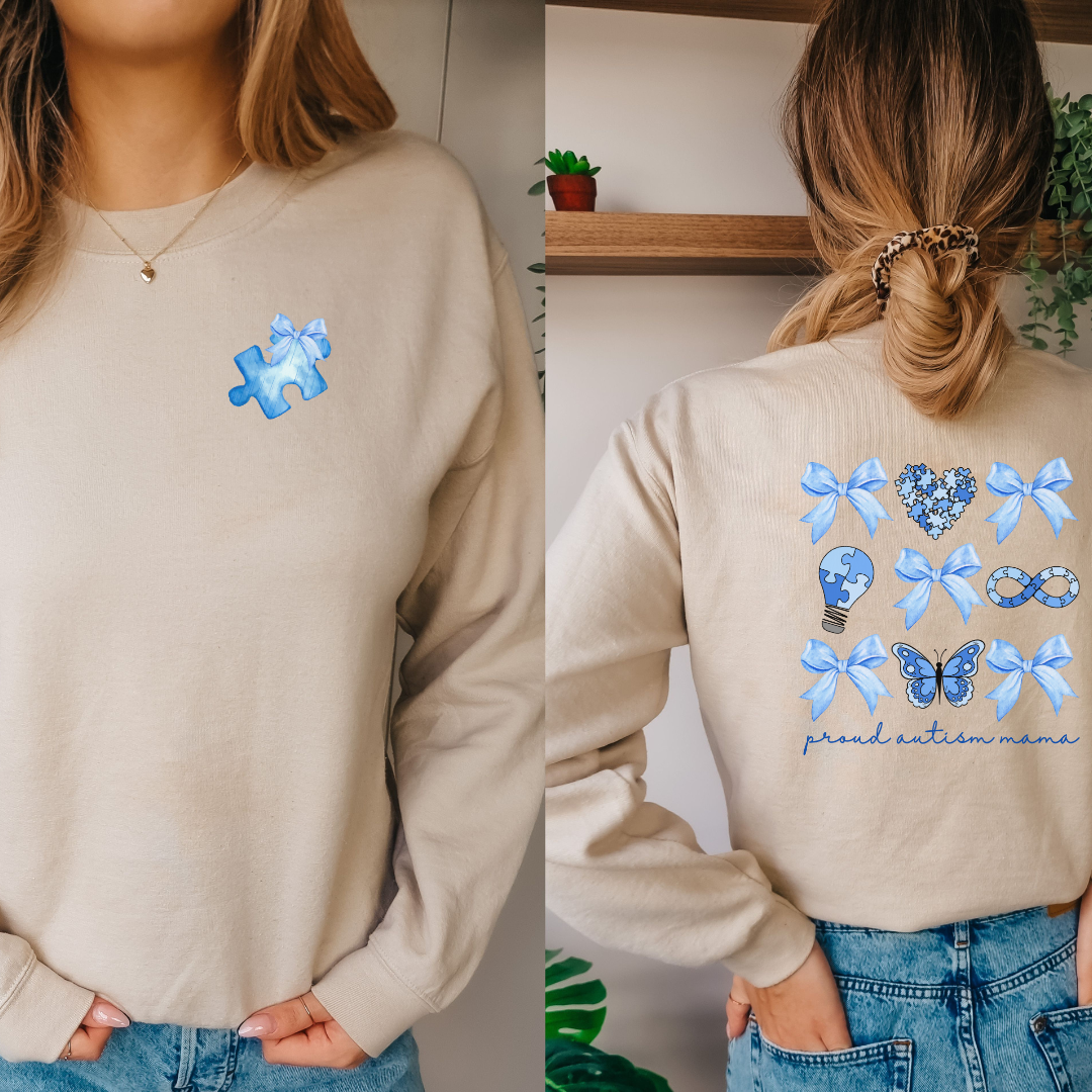 Sand colored sweatshirt with a soft blue puzzle piece  with a bow on the left chest. On the back blue bows and Autism pieces in a Coquette style that says proud autism mama